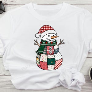 Op de afbeelding: Wit T-shirt met een grillig sneeuwpopontwerp. De sneeuwpop draagt een rood-witte geruite hoed, een groene sjaal en een patchwork lichaam met verschillende patronen en kleuren. Hij heeft een wortelneus en takarmen.
