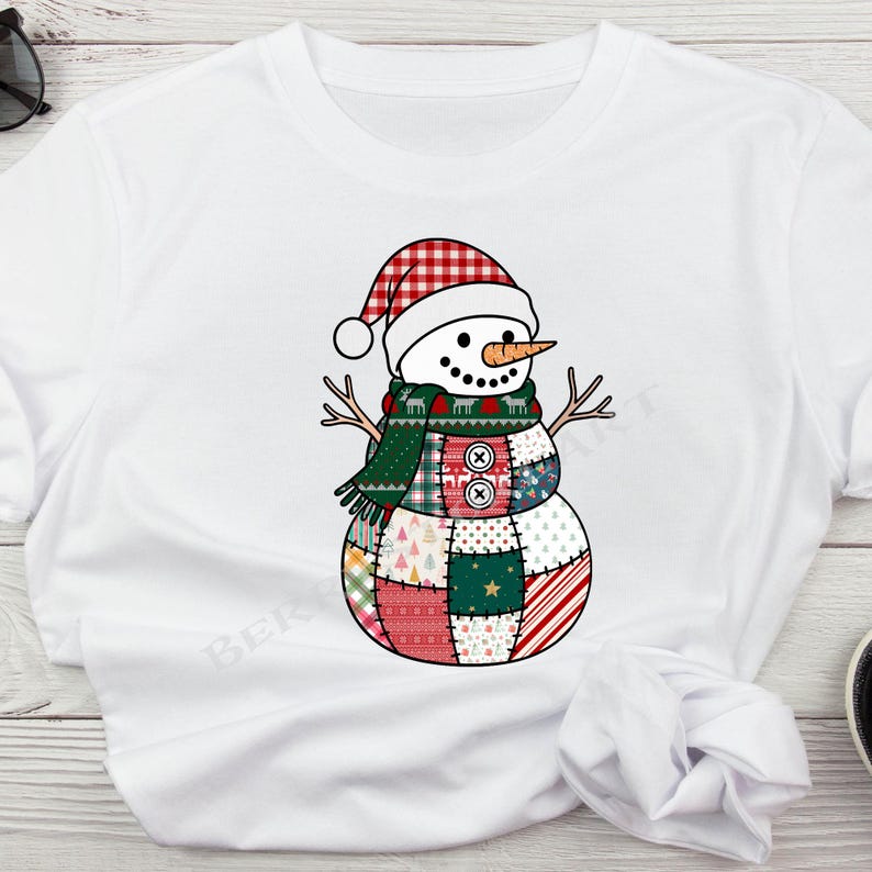 Op de afbeelding: Wit T-shirt met een grillig sneeuwpopontwerp. De sneeuwpop draagt een rood-witte geruite hoed, een groene sjaal en een patchwork lichaam met verschillende patronen en kleuren. Hij heeft een wortelneus en takarmen.
