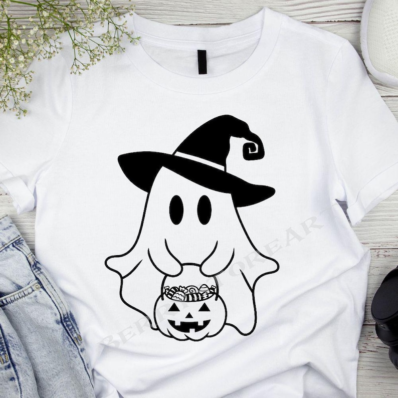 May include: White t-shirt with a black and white illustration of a ghost wearing a witch hat and holding a pumpkin filled with candy.