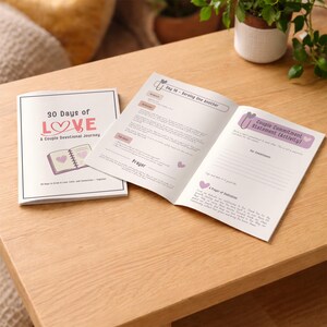 May include: A devotional book titled "30 Days of Love: A Couple Devotional Journey" is open on a wooden table. The open pages display text and prompts for reflection. The cover features the title in pink and a heart illustration.