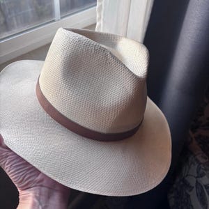 May include: A cream-colored woven straw fedora hat featuring a pinched crown and a wide brim. The hat is accented with a smooth, solid brown leather band encircling the base of the crown, providing a distinct contrast to the light-colored straw texture.