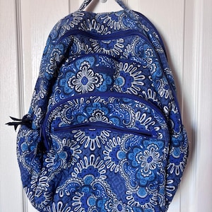 May include: A quilted fabric backpack featuring a vibrant blue and white paisley floral pattern. The bag includes a top handle, two main zippered compartments, and a smaller front zippered pocket, all finished with matching blue zipper pulls.
