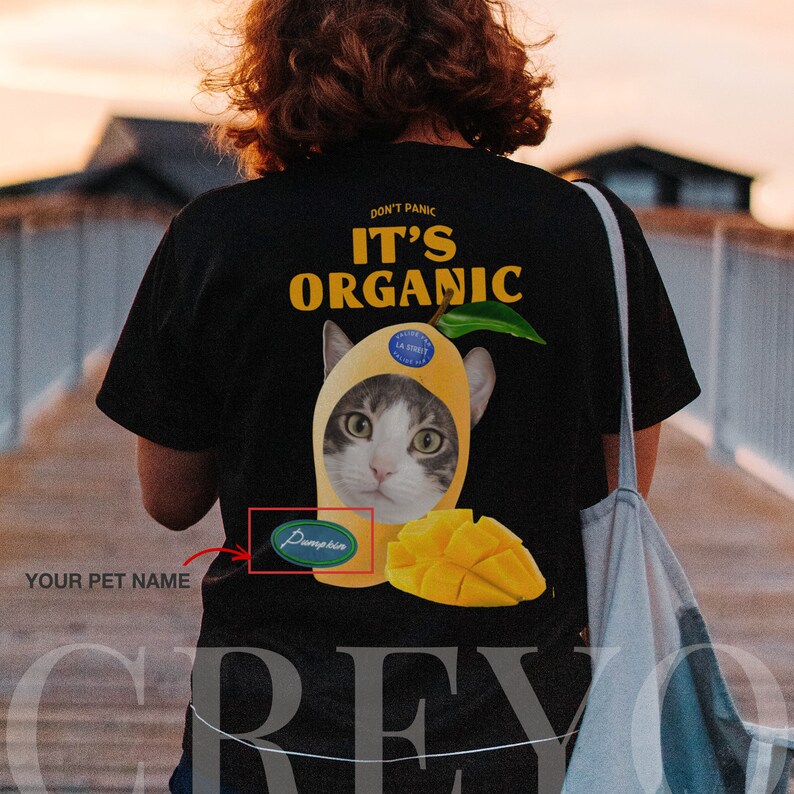 May include: Black t-shirt with a yellow mango design and a cat's face inside. The text "Don't Panic It's Organic" is printed on the shirt. A green sticker with the word "Pumpkin" is on the shirt.