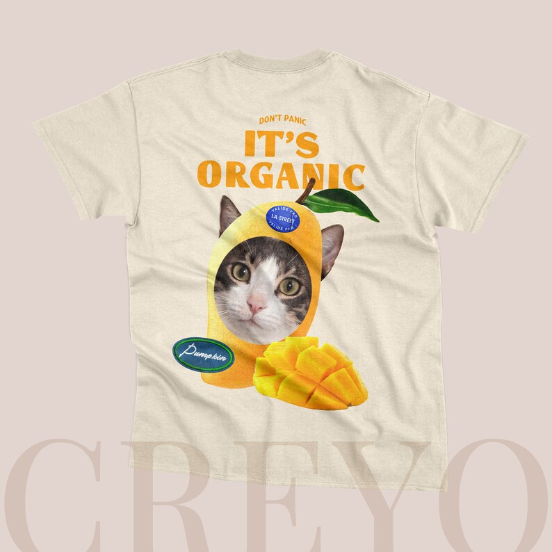 May include: A beige t-shirt with the text "Don't Panic It's Organic" printed on the back. A cat's head is peeking out of a mango with a sticker that says "Valeur La Source La Source" on it. The mango is on top of a smaller mango slice.