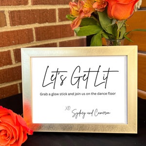 May include: A rectangular gold-toned picture frame containing a white sign with black text that reads Let's Get Lit, Grab a glow stick and join us on the dance floor, and XO Sydney and Cameron, displayed with orange flowers.