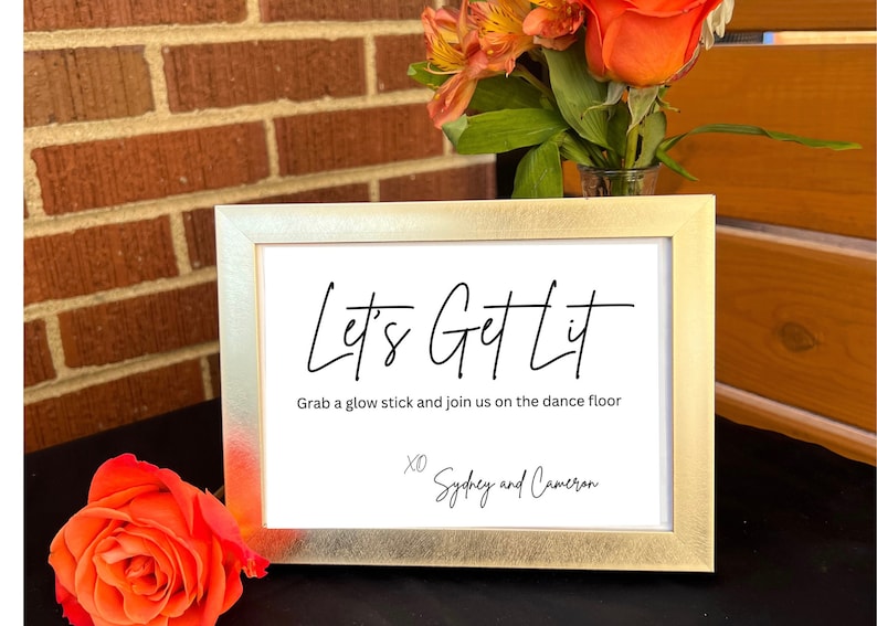 May include: A rectangular gold-toned picture frame containing a white sign with black text that reads Let's Get Lit, Grab a glow stick and join us on the dance floor, and XO Sydney and Cameron, displayed with orange flowers.