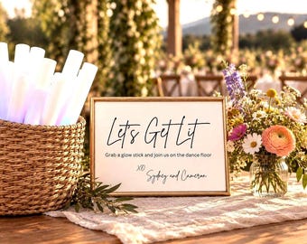 May include: A rectangular gold-framed sign with black text reading Let's Get Lit, Grab a glow stick and join us on the dance floor, XO Sydney and Cameron, next to a woven basket filled with white glow sticks and a glass vase of colorful flowers.