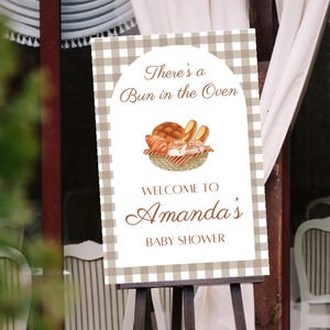May include: A sign on an easel with a gingham pattern background. The sign reads "There's a Bun in the Oven" above a watercolor illustration of baked goods. Below, it says "Welcome to Amanda's Baby Shower."