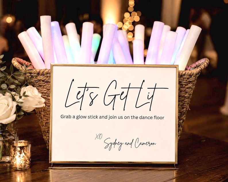 May include: A rectangular gold-framed sign with white background and black text reading Let's Get Lit, Grab a glow stick and join us on the dance floor, XO Sydney and Cameron, placed in front of a woven basket filled with glowing LED foam sticks.