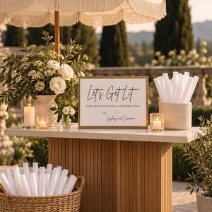 May include: A rectangular sign with a gold frame displays the text Let's Get Lit, Grab a glow stick and join us on the dance floor, XO, Sydney and Cameron. It sits on a table with white candles, flowers, and white glow sticks in containers.