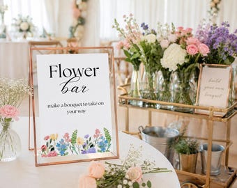 以下が含まれることがあります： 銅色のフレームに入った長方形の看板。Flower barおよびmake a bouquet to take on your wayという文字が書かれ、下部にはカラフルな花のイラストが描かれています。白いテーブルの上に置かれ、周囲には切り花が添えられています。