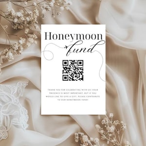 Puede incluir: Una tarjeta blanca con las palabras "Honeymoon Fund" en elegante escritura negra, con un código QR y un mensaje. La tarjeta está colocada sobre un fondo de tela suave de tonos neutros y delicados acentos florales.