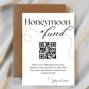 Puede incluir: Una tarjeta blanca con las palabras "Honeymoon Fund" en elegante escritura negra, acompañada de un código QR y un mensaje. La tarjeta se combina con un sobre marrón, sobre un fondo suave y blanquecino.