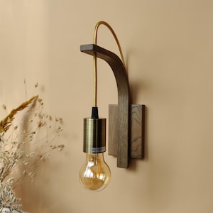 Può includere: Un'applique da parete in legno con un braccio curvo e un apparecchio di illuminazione in ottone. L'applique è montata su una parete e ha un cavo dorato.