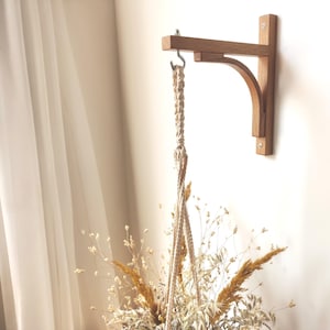 May include: A light brown wooden wall bracket with a curved support arm and a silver metal hook attached to the end. A cream-coloured macrame cord hanger is suspended from the hook, holding a bouquet of dried grasses and flowers in neutral tones.