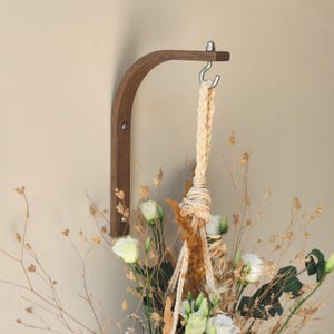 May include: A dark wooden wall-mounted plant hanger with a curved arm and a metal hook. The hanger is designed to hold a hanging plant or other decorative items. A woven rope is attached to the hook. The hanger is mounted on a neutral-coloured wall. The wood is stained.