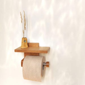 May include: Wooden toilet paper holder with a small shelf and a decorative vase holding dried grass. The holder is made of light brown wood and includes a roll of tan toilet paper. The vase is a light yellow color with a small window design.