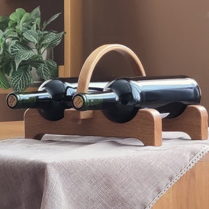 May include: A wooden wine rack holding two dark glass wine bottles. The rack has a curved handle and a base designed to cradle the bottles. The bottles have green necks and gold labels. The rack is made of light brown wood.