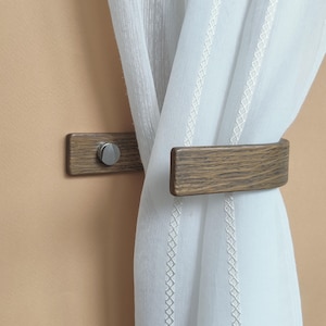 May include: A wooden curtain tieback with a brushed nickel screw. The tieback is attached to a wall and holds a white sheer curtain.