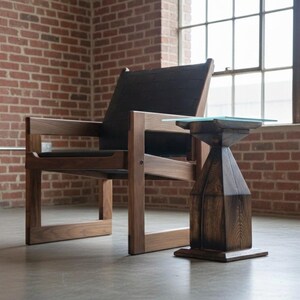 May include: A modern wooden armchair with a dark seat and back, paired with a small side table. The table has a geometric wooden base and a square glass top. The furniture is set against a brick wall and a window.