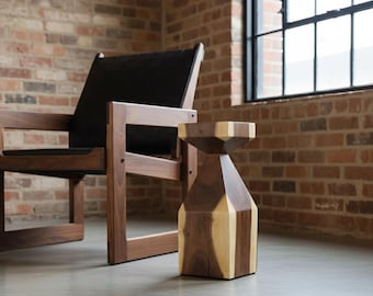 May include: A wooden side table with a geometric design, featuring alternating sections of light and dark wood. The table is approximately 18 inches tall and stands on a concrete floor. A wooden chair with a black seat is in the background.
