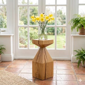 May include: A wooden side table with a geometric design, supporting a clear glass vase filled with yellow daffodils. The table is made of light-colored wood and stands on a terracotta tile floor. The room has large windows and natural light.