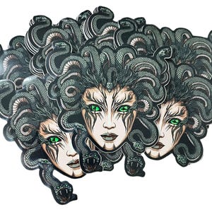 Könnte beinhalten: Ein Stapel Aufkleber mit einer grünen und schwarzen Illustration von Medusa, einer mythischen griechischen Figur mit Schlangenhaaren. Ihr Gesicht ist mit einem grimmigen Ausdruck und durchdringenden grünen Augen dargestellt.