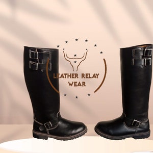 Op de afbeelding: Een paar zwarte leren laarzen die tot op de knie komen met dubbele gespen en een bandje over de enkel. De laarzen hebben een dikke hak en een afgeronde neus. De laarzen staan op een wit platform. De tekst "LEATHER RELAY WEAR" is zichtbaar op de achtergrond.