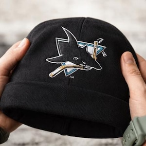 Puede incluir: Gorro de punto negro con el logotipo de los San Jose Sharks. El logotipo representa un tiburón con un palo de hockey y un triángulo con un contorno verde azulado y blanco. El gorro está sujeto por dos manos.