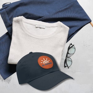 Puede incluir: Una gorra de béisbol azul marino con un parche de cuero marrón que presenta un sol y el texto "Be Sunshine".