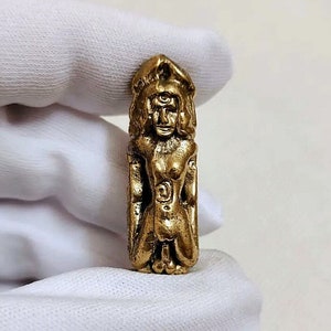 Könnte beinhalten: Ein goldfarbener Metallanhänger, der eine kniende Figur mit einem dritten Auge auf der Stirn darstellt. Die Figur ist von komplizierten Details und Symbolen umgeben.