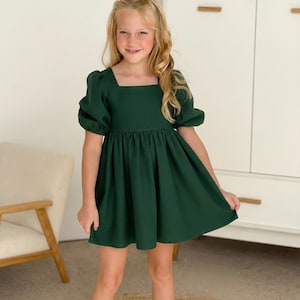 May include: A dark forest green dress featuring a square neckline, short puff sleeves with gathered detailing, and a gathered waistline that flows into a flared skirt. The garment is constructed from a solid-colored fabric with a visible texture.