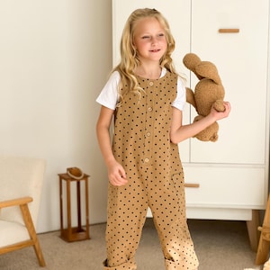 以下が含まれることがあります： 白いTシャツの上に、ベージュのドット柄のジャンプスーツを着た子供。白いサンダルを履いています。ジャンプスーツは黒い水玉模様で、ボタン留めです。子供は茶色のぬいぐるみを持っています。