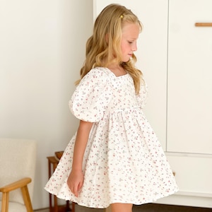 May include: A white floral dress with a pattern of small pink and red flowers. The dress has a square neckline and short, puffed sleeves. The skirt flares from the waist. The dress is worn by a young person.
