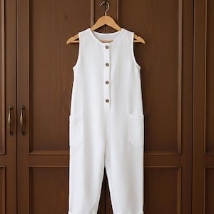 Puede incluir: Un mono blanco sin mangas con botones en la parte delantera y dos bolsillos delanteros cuelga de una percha de madera. El mono tiene un corte holgado con perneras recortadas. Los botones son de color marrón claro. El fondo es una puerta de madera marrón oscuro.