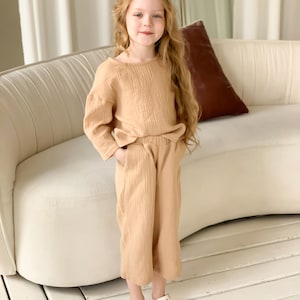 May include: A tan two-piece outfit for children. The set includes a long-sleeved top and cropped pants. The top has a round neckline and a bow detail at the waist. The pants have pockets. The outfit is made of a textured fabric.