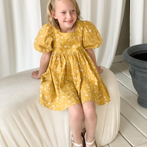 Könnte beinhalten: Ein senfgelbes Kleid mit weißem Blumenmuster und Puffärmeln. Das Kleid hat einen quadratischen Ausschnitt und einen gerafften Rock. Das Mädchen trägt weiße Sandalen mit braunen Sohlen. Das Kleid ist knielang.