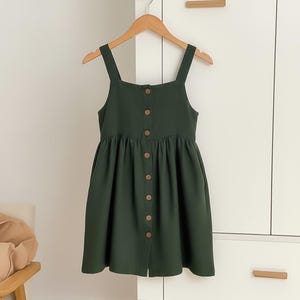 Puede incluir: Vestido verde oscuro con botones en la parte delantera y tirantes anchos. El vestido está colgado de una percha de madera sobre un fondo blanco. El vestido tiene una falda fruncida y botones de madera.