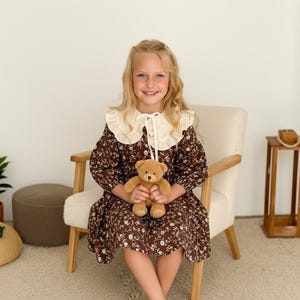 May include: A young person wearing a brown floral dress with a cream-colored Peter Pan collar, holding a teddy bear. The dress has long sleeves and a knee-length hem. The person is seated in a light-colored chair.