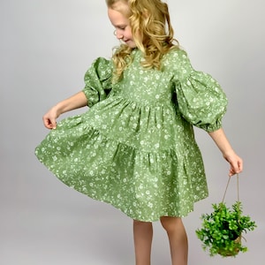 Pode incluir: Uma menina pequena vestindo um vestido verde floral com mangas bufantes. Ela está segurando uma pequena planta em vaso.
