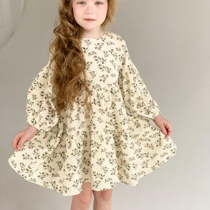 May include: A cream-colored dress with a floral pattern featuring green leaves and red berries. The dress has long, puffy sleeves and a tiered skirt. The girl is wearing white sandals.