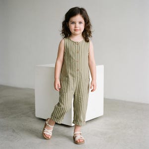 May include: A sleeveless olive green jumpsuit featuring vertical white stripes, a round neckline, a front button placket with four visible buttons, and two front patch pockets. The pant legs are cuffed at the bottom, paired with white sandals.