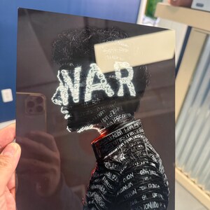 May include: A black and white art print featuring a profile of a person with the word "WAR" in white neon-style lettering. The person's form is detailed with text. The print is held by a hand.