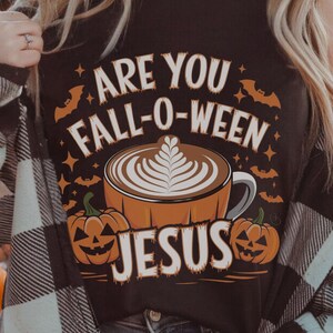 Pode incluir: Camiseta preta com um design gráfico que apresenta uma abóbora, uma xícara de café com arte latte e o texto "Are you Fall-O-Ween Jesus".