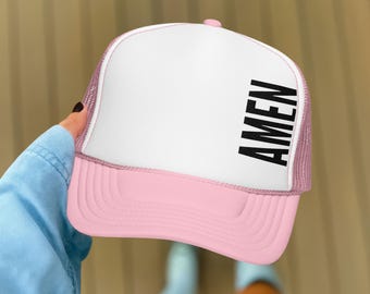 May include: A pink and white trucker hat with the word "AMEN" printed vertically in black on the front. The hat has a mesh back and a curved brim. The hat is being held up by a person.