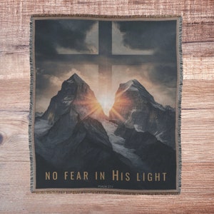 Pode incluir: Uma manta de tecido tecido com uma cena de montanha e uma cruz. As montanhas são silhuetas contra um céu de pôr do sol. O texto "NO FEAR IN HIS LIGHT" e "PSALM 27:1" está impresso na manta.