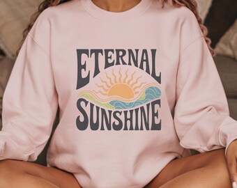 May include: A pink sweatshirt with the text "Eternal Sunshine" in a retro font. The text is surrounded by a graphic of a sun setting over waves.