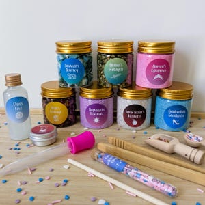 May include: A collection of jars and bottles with gold lids and labels, including text such as "Elsa's Love" and "Jasmine's Bravery." The jars contain colourful contents, with wooden scoops and a dropper. The theme appears to be inspired by Disney characters.