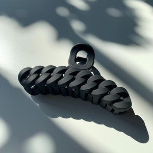 May include: A matte black hair claw clip with a chain link design. The clip is made of a solid, dark material and is designed to hold hair in place. The claw clip is on a white surface with shadows.