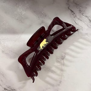 May include: A burgundy hair claw clip with a gold-colored cat silhouette. The clip is made of glossy plastic and has a claw-like design with multiple teeth for securing hair. The clip is approximately 4 inches long.
